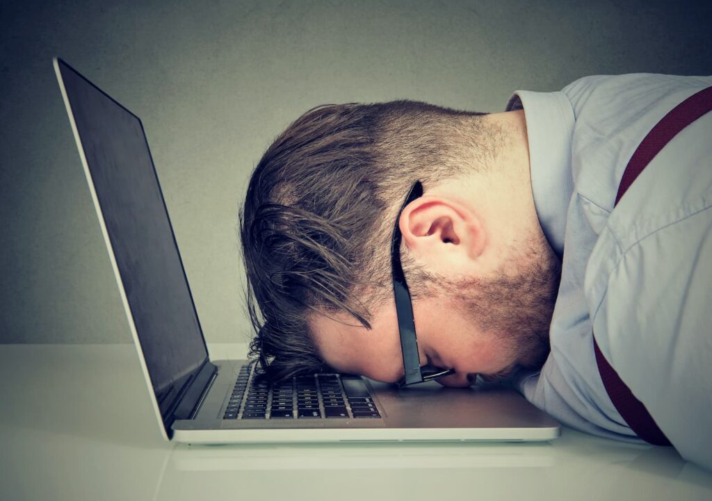 Frustrated or exhausted man with glasses resting his head face-down on a laptop keyboard, conveying stress or burnout from work or technology.