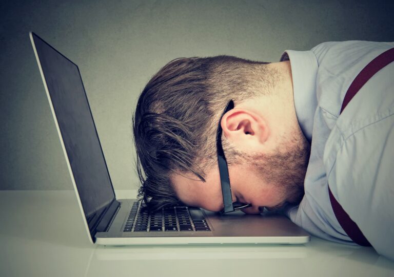 Frustrated or exhausted man with glasses resting his head face-down on a laptop keyboard, conveying stress or burnout from work or technology.