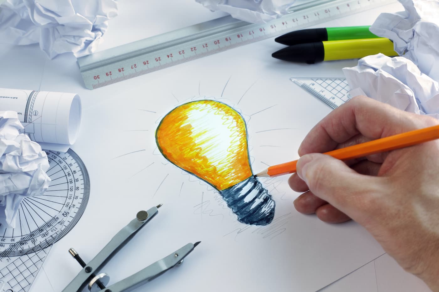 A hand drawing a colorful light bulb with an orange pencil on a sheet of paper, surrounded by crumpled paper balls, drafting tools, and markers—symbolizing the creative process and brainstorming ideas.