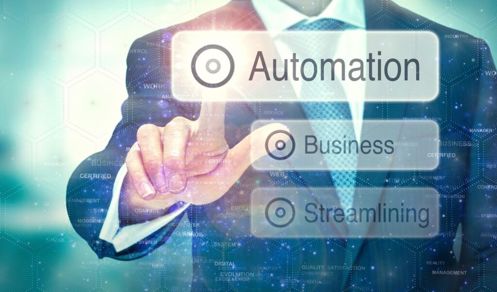 A businessman in a suit is touching a virtual screen, selecting the word “Automation” from a digital interface. Below it are the words “Business” and “Streamlining.” The background features abstract digital elements, hexagonal patterns, and tech-related words, symbolizing technology integration in modern business processes.