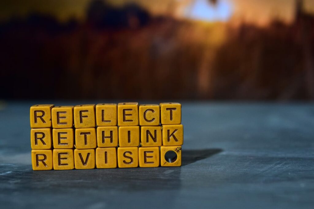Yellow letter blocks stacked on a table spell out the words “REFLECT,” “RETHINK,” and “REVISE” in three rows. The background is softly blurred with warm, dark tones, drawing focus to the motivational words in the foreground.
