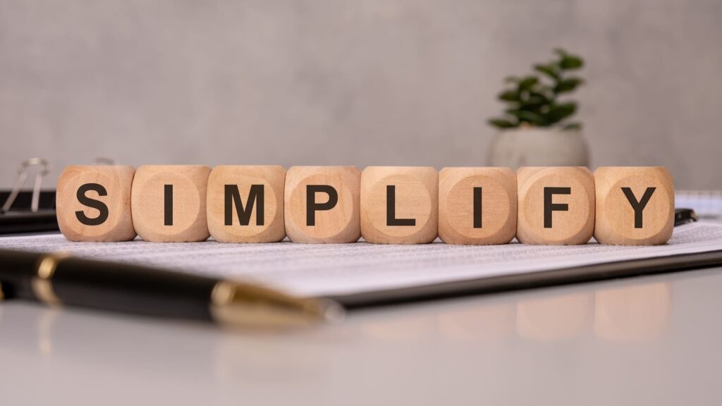 Wooden blocks on a desk spell out the word "SIMPLIFY" in bold black letters. The blocks are arranged in a neat row atop a document, with a pen and small potted plant in the background, suggesting a minimalist or organized workspace.
