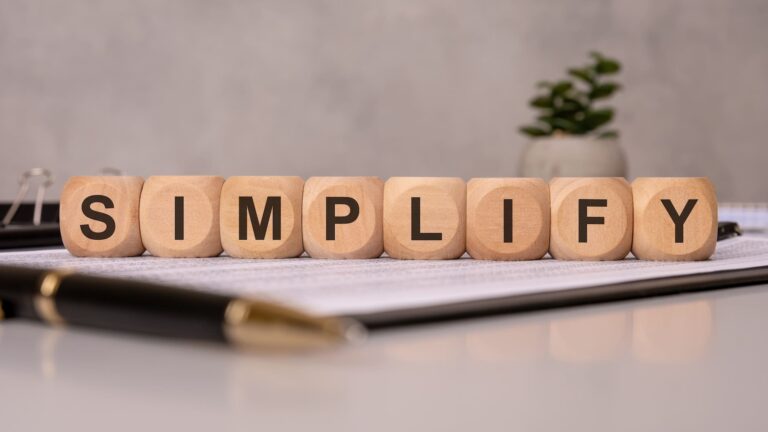 Wooden blocks on a desk spell out the word "SIMPLIFY" in bold black letters. The blocks are arranged in a neat row atop a document, with a pen and small potted plant in the background, suggesting a minimalist or organized workspace.