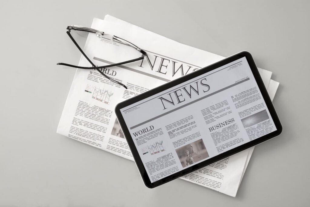 A tablet displaying a digital newspaper lies on top of a printed newspaper with similar layout and content. A pair of reading glasses rests on the paper, highlighting the contrast between traditional and digital news formats.