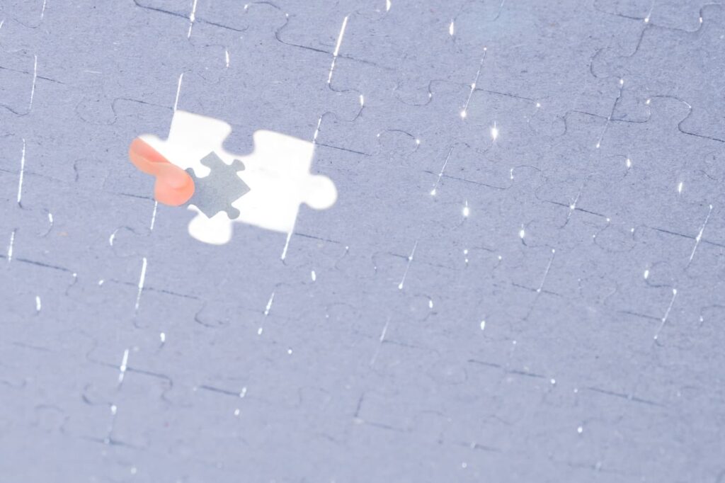 A close-up of a nearly completed jigsaw puzzle with one piece missing. A person’s hand with a peach-colored nail is holding the final puzzle piece above the empty spot, ready to place it. Light reflects off the glossy surface of the puzzle, creating small sparkles.