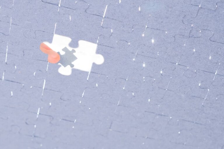 A close-up of a nearly completed jigsaw puzzle with one piece missing. A person’s hand with a peach-colored nail is holding the final puzzle piece above the empty spot, ready to place it. Light reflects off the glossy surface of the puzzle, creating small sparkles.