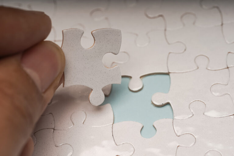 A close-up of a nearly completed jigsaw puzzle with one piece missing. A person’s hand is holding the final puzzle piece above the empty spot, ready to place it. Light reflects off the glossy surface of the puzzle, creating small sparkles.