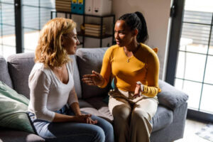 Portrait of two diverse mid adult female friends sitting on the sofa, face to face, having a sincere conversation. One consoling the other.