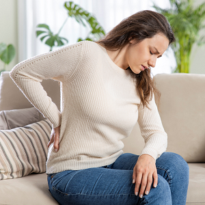 woman sitting on a couch looking down with her hand on her back seeming to be in pain