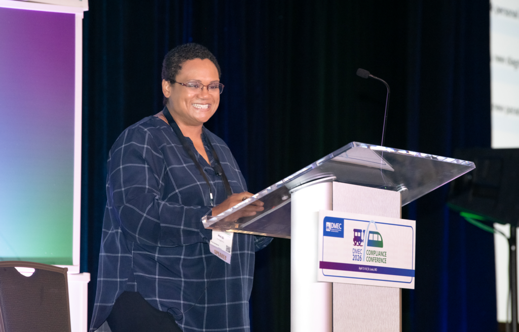 A smiling speaker presents at a podium during a DMEC conference session.